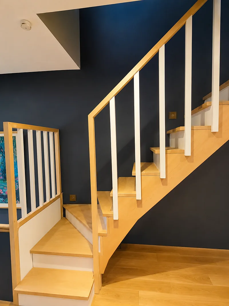 Escalier quart tournant en bois clair avec garde-corps à barreaux blancs, devant un mur bleu nuit