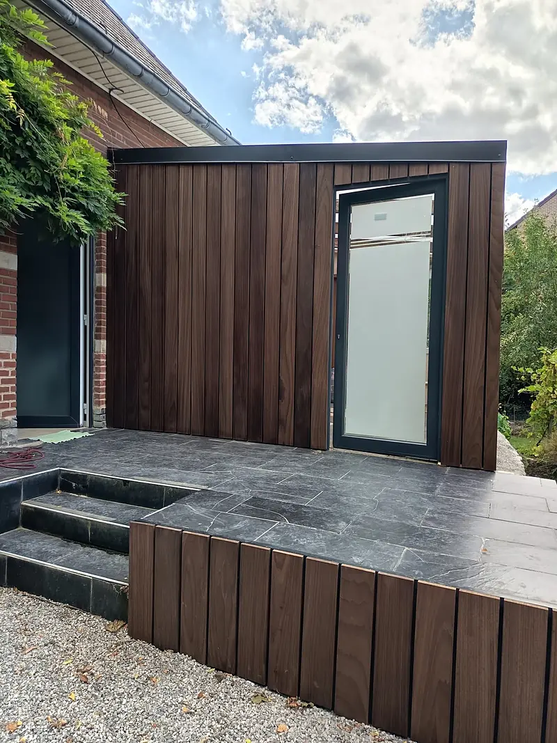 Véranda à bardage bois avec porte vitrée, donnant sur une terrasse en pierre